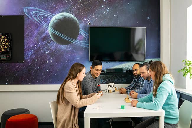 Das Foto zeigt eine Gruppe von Mitarbeiterinnen und Mitarbeitern im farbenfrohen Pausenraum am Tisch sitzend. Den Pausenraum schmückt eine Weltraum-Tapete, bunte Hocker, eine Dart-Scheibe und ein Spielekonsole. Die Mitarbeiter interagieren miteinander.