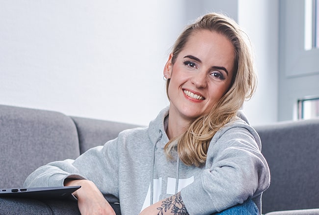 Eine junge Frau namens Stephi sitzt in einer gemütlichen Sitzecke auf dem Boden bei BROCKHAUS AG. Sie hat einen Laptop vor sich aufgestellt und trägt einen grauen Hoodie.